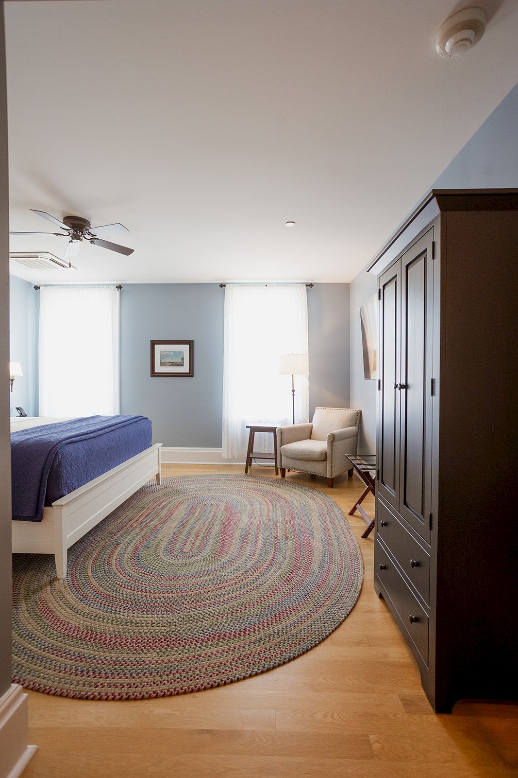 A bright bedroom with a bed, round patterned rug, ceiling fans, an armchair by a lamp, and a dark wood wardrobe dresser.