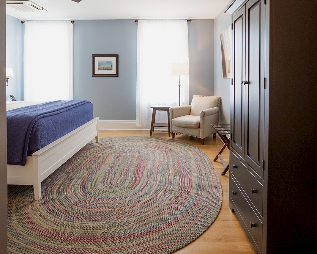 A bright bedroom with a bed, round patterned rug, ceiling fans, an armchair by a lamp, and a dark wood wardrobe dresser.