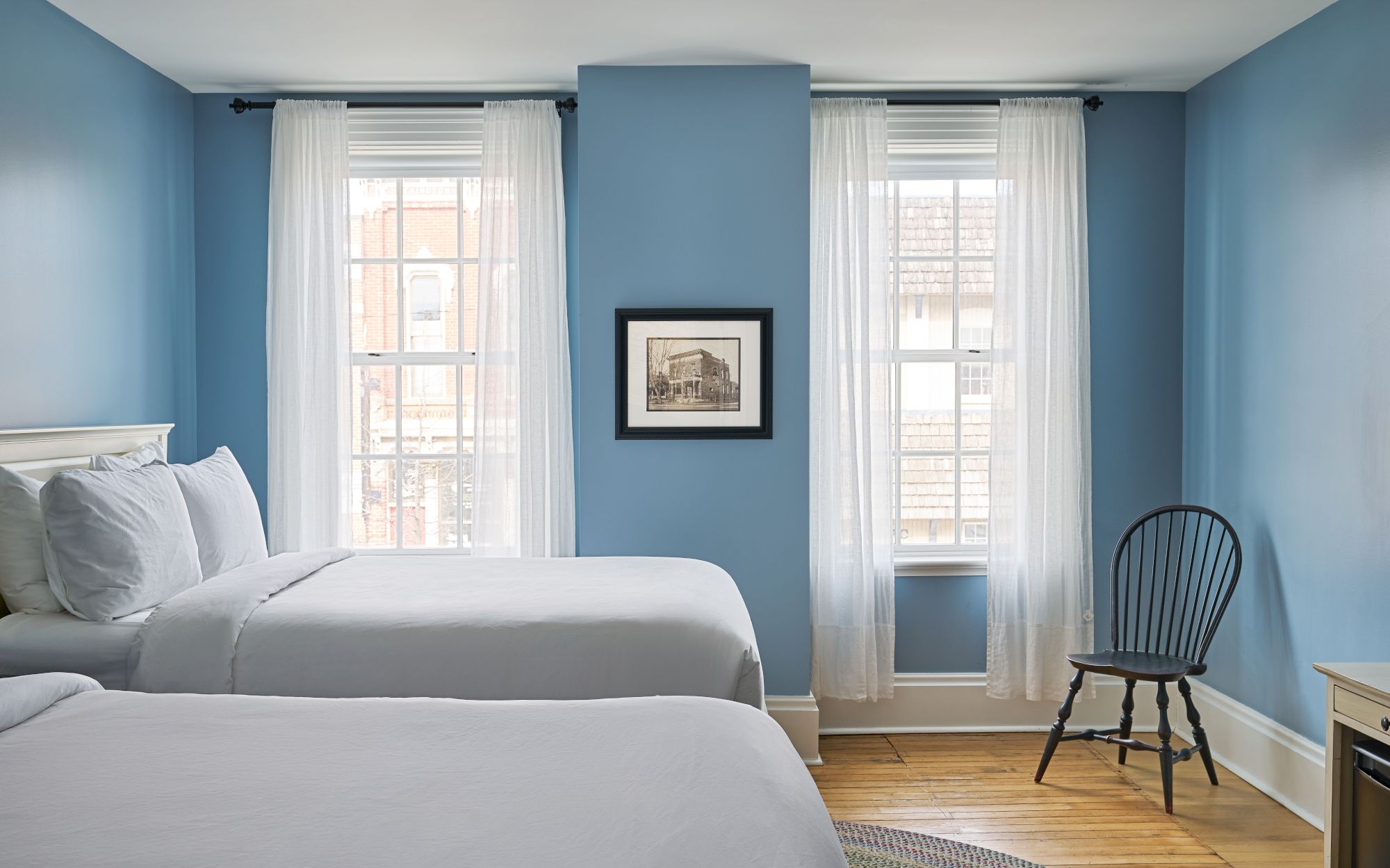 A bedroom with two beds, blue walls, white curtains, a framed picture, and a wooden chair by the window. The room is bright and simple.