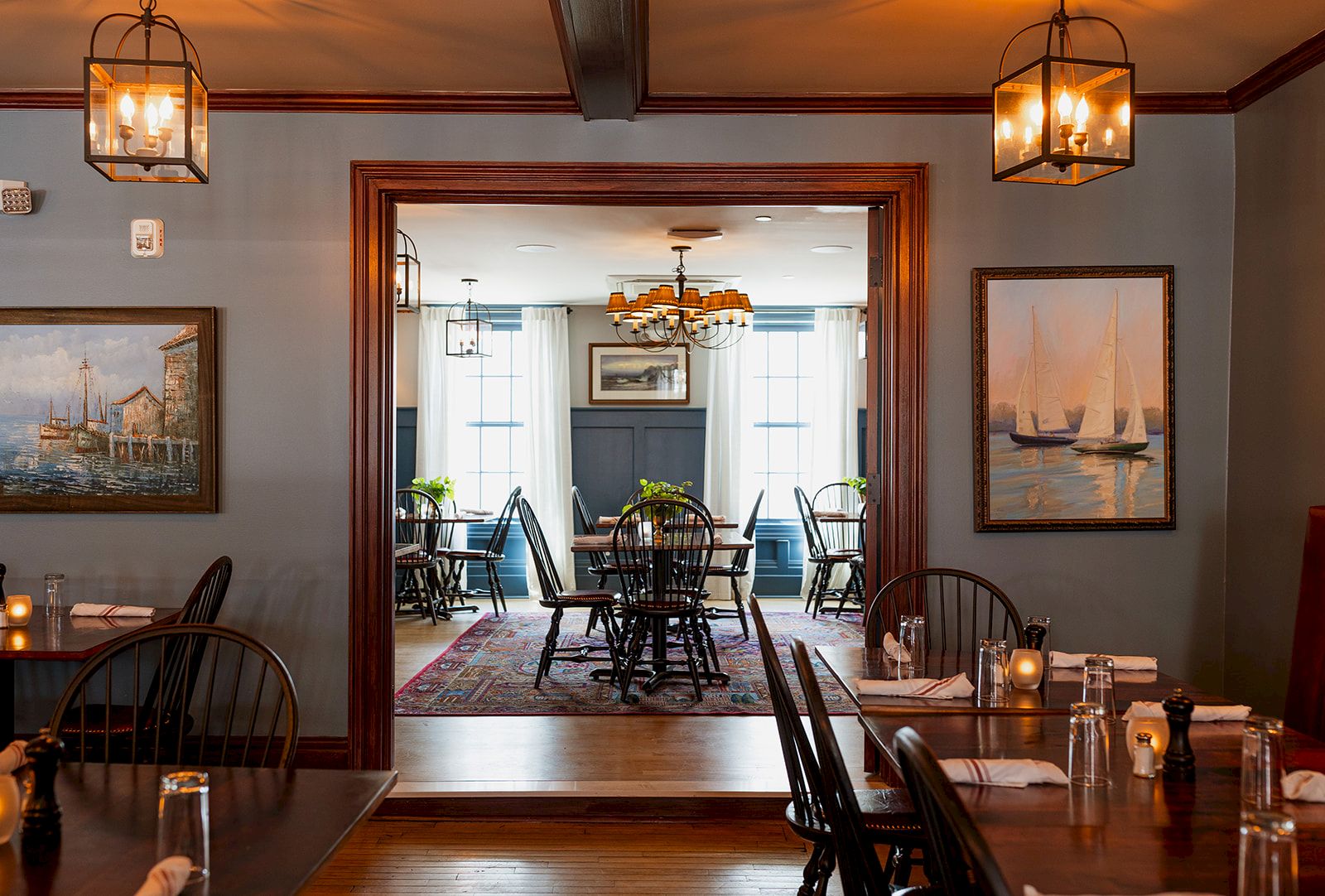 Warm dining room with wood tables, mismatched chairs, chandeliers, and framed sailboat paintings visible through an open doorway.