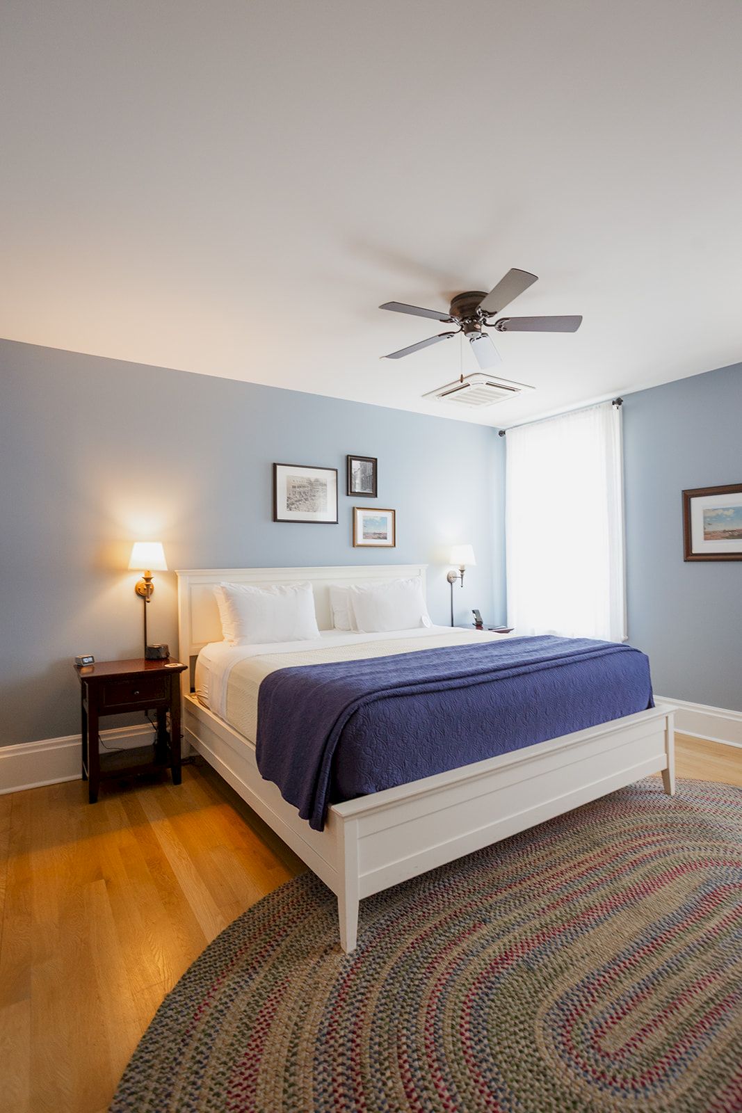 A tidy bedroom with a white bed, bedside lamps, a patterned rug, and a ceiling fan above it.